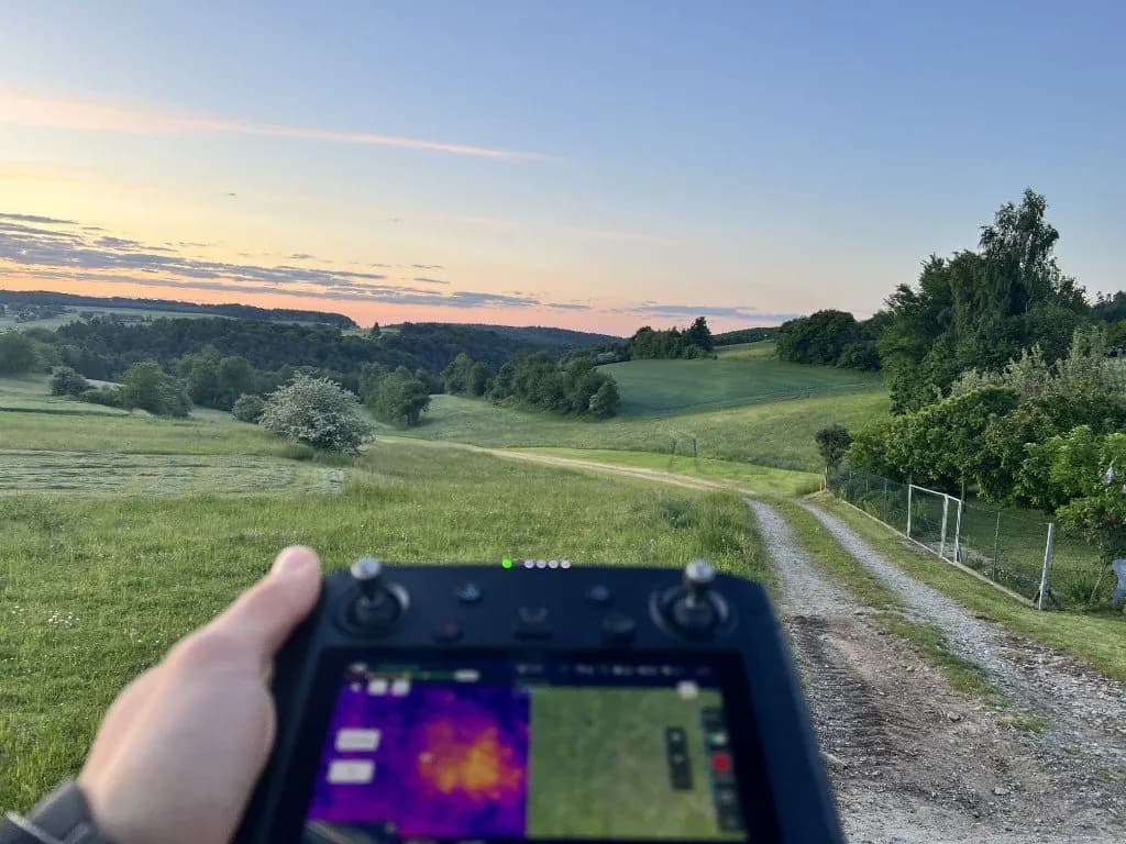 Drone controller with thermal and visual feeds over a green field at dawn — fawn-rescue field documentation.
